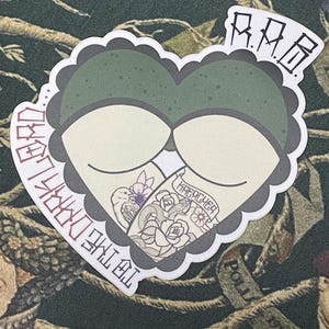 May include: A green heart-shaped sticker with a white outline and the text "RIP" at the top. The heart is filled with a white background and a black outline. The text "The End" is written in red on the left side of the heart, and the text "Forever" is written in black on the right side of the heart. The heart is decorated with a black and white tattoo design of a snake, flowers, and a bird.