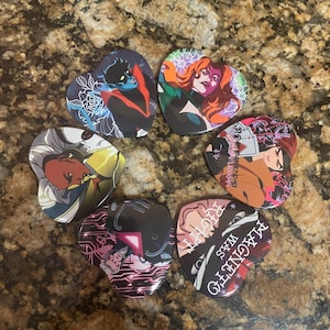 May include: A collection of heart-shaped pins featuring comic book characters. Each pin displays a different character with vibrant colors and unique designs. The pins are arranged in a circular pattern on a textured surface.