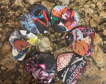 May include: A selection of heart-shaped pins showcasing comic book characters. Each pin features a different character with vivid colours and distinctive designs. The pins are arranged in a circular pattern on a textured surface.