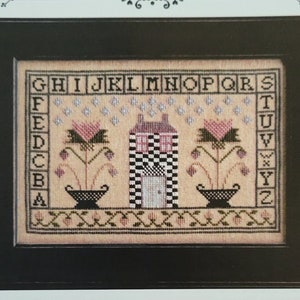 May include: Cross stitch pattern for a counted thread embroidery project titled "Little Brick House". The design features a small house with a black and white checkered pattern, two pink flowers in pots, and a border of small flowers. The pattern includes an alphabet chart.