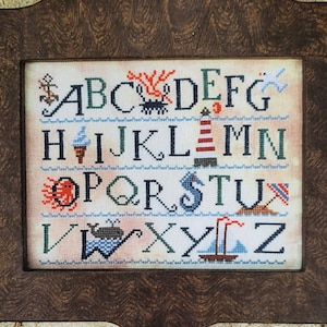 May include: A framed cross-stitch sampler titled "Sampler of the Sea" with a nautical theme. The sampler features the alphabet with sea-related images. The frame is dark brown, and the background is a sandy color. The text "Carriage House Samplings" is at the top and bottom.