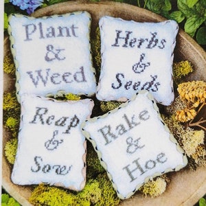 May include: Four white fabric squares with embroidered text: "Plant & Weed", "Herbs & Seeds", "Reap & Sow", and "Rake & Hoe". The squares are arranged in a bowl with green moss and flowers.