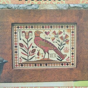 May include: A cross-stitch pattern featuring a stylized bird with a red, orange, and yellow design. The bird is surrounded by flowers and other decorative elements. The pattern is titled "Frederick" and is from Carriage House Samplings.