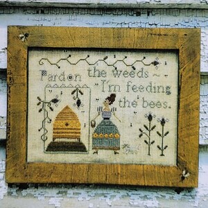 May include: A framed cross-stitch sampler with the text "Pardon the weeds ~ I'm feeding the bees." The artwork features a beehive, a woman, and floral elements. The frame is a rustic, light brown color. The words "Notforgotten Farm 1887" are at the top.