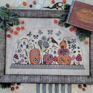 May include: Cross stitch pattern for an autumn cloche with pumpkins, butterflies, and a bird. The pattern is framed and surrounded by fall-themed decorations, including buttons, yarn, and small pumpkins.