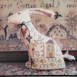 May include: A cross-stitch pattern for a sampler featuring a white rabbit with a red heart and a small house. The pattern is titled "Samplar Hare" and is by La-D-Da designs for counted thread.