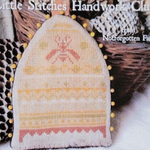 May include: A cross-stitch pattern of a beehive with a bee inside, titled "Queen Bee Needle Poke" from the Little Stitches Handwork Club. The pattern is displayed on a piece of fabric with yellow and orange accents.