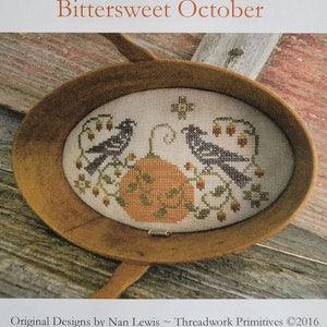 May include: A cross-stitch pattern titled Bittersweet October by Threadwork Primitives. The design features two birds flanking a central orange pumpkin surrounded by vines and berries, displayed inside an oval wooden bowl on a rustic surface.