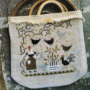 May include: A cross-stitch pattern sampler with a beige background and a design of a small house, a tree, and birds. The sampler is framed in a wooden hoop and is hanging on a wooden surface. The text "Chickweed" is printed below the sampler.