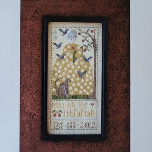 May include: Cross stitch pattern featuring a woman in a yellow dress with white polka dots, holding a book. The woman is surrounded by birds and a cat. The text "Marion the Librarian 12-11-2002" is stitched below the woman. The pattern is framed in black.