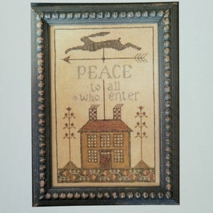 May include: Framed cross-stitch art with the text "Peace House" and "Peace to all who enter." The artwork features a house, a rabbit weathervane, and floral accents. The frame is dark with a beaded edge. The bottom text reads "La-D-Da designs for counted thread."