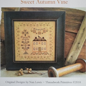 Op de afbeelding: Een ingelijste kruissteeksampler met de titel "Sweet Autumn Vine" met een huis- en bloemenontwerp. De sampler staat in een zwart frame en bevat de tekst "Autumn". Een houten spoel en decoratieve elementen staan op de voorgrond. De afbeelding bevat de tekst "Threadwork Primitives".