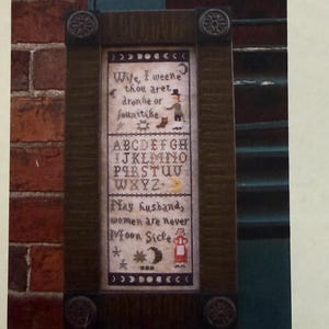 May include: A framed cross-stitch sampler with the text "Moon Sicke" above it. The sampler features an alphabet, a verse, and decorative elements. A smiling moon graphic is to the right. The Primitive Needle logo is at the top.