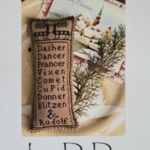 May include: A Christmas-themed design featuring a stitched pillow with reindeer names, a Christmas card, and evergreen sprigs. The pillow is tan with black stitching and the names of Santa's reindeer. The card says "Merry Christmas". The text "And Rudolf" and "La-D-Da" are also visible.