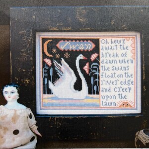 May include: A cross-stitch pattern featuring a white swan with a black eye and a black beak, swimming in a blue pond with green reeds. The pattern is framed in a black frame and includes the text "Swan" and a poem about swans. The image is from Carriage House Samplings.