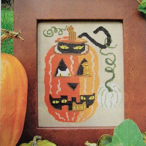May include: Cross stitch pattern for a pumpkin with cat eyes. The pattern is called "Cats' Eyes" and is from Carriage House Samplings. The image shows a framed cross stitch of the pattern, with a real pumpkin in the foreground.