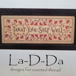 May include: A framed cross-stitch design with the text "TREAT YOUR SELF WELL" in a brown frame. The design is by La-D-Da, designs for counted thread.