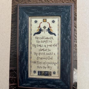 May include: A framed cross-stitch sampler titled "The Dampy Sod" with a dark blue frame. The sampler features a central design with two birds and text. The top of the image has a vintage image and the words "The Primitive Needle".