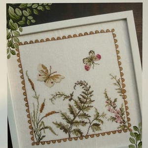 Puede incluir: Un diseño de punto de cruz enmarcado con una mariposa y elementos florales. La obra de arte está en un marco blanco con un borde decorativo. El texto "Flutterby" está en la parte inferior, con "Mojo Stitches" y "Needlework Designs & Delights" en la parte superior.
