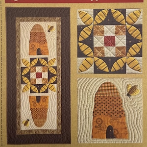 May include: A quilting pattern titled Gathering Honey by Timeless Traditions. The cover features a finished quilt design with bee skeps, bees, and floral blocks in brown, tan, and yellow. Text includes a quote and finished size 21 x 45 inches.