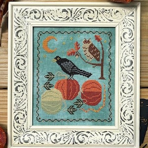 May include: Framed cross-stitch art featuring a black crow perched on orange pumpkins. A brown owl sits on a tree branch, with a crescent moon in the sky. The frame is white with ornate detailing, perfect for fall decor.