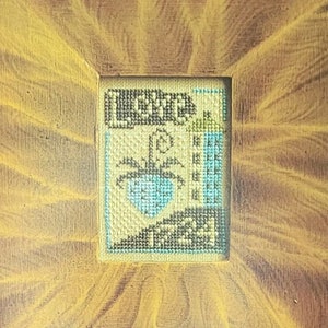 May include: A cross-stitch pattern sampler with a vintage-style design. The pattern features a small house with a blue roof and a tree. The text "Love" is printed at the top of the image. The text "The Virtue Sampler Series" is printed below the image. The text "Charity Faith Honor Hope Love" is printed below the series title. The text "Carriage House Samplings" is printed below the virtue list. The text "www.CarriageHouseSamplings.com" is printed below the company name.