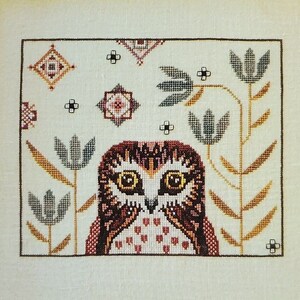 May include: A counted cross stitch pattern featuring an owl with brown, pink, and white feathers, surrounded by stylized plants and flowers. The pattern is titled "Oona Owl" and is by The Artsy Housewife.