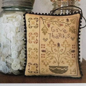 Op de afbeelding: Een kruissteek kussen met de woorden "Give Thanks" en het jaar 2014. Het kussen is beige met bruine en groene steken en een zwarte pompom afwerking. Een glazen pot gevuld met knopen en een andere met gedroogde bessen staan op de achtergrond.