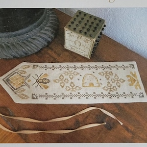 Op de afbeelding: Een witte linnen bladwijzer met een bij en honingraat ontwerp in gouden en bruine draad. De bladwijzer is vastgebonden met een crèmekleurig lint. De tekst "A Bee's Gathering" is gedrukt in gouden letters op een witte achtergrond. De tekst "Original Designs by Nan Lewis - Threadwork Primitives ©2013" is gedrukt in zwarte letters op een witte achtergrond. De tekst "Email: threadworkprimitives@verizon.net" is gedrukt in zwarte letters op een witte achtergrond.