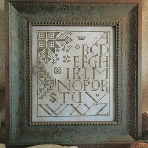May include: A framed cross-stitch sampler featuring the alphabet in a dark thread on a light fabric background. The text reads Threadwork Primitives, Quaker Block Print, and Original Designs by Nan Lewis ~ Threadwork Primitives ©2021.