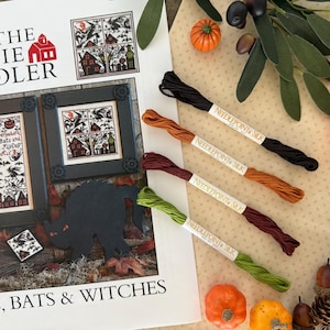 May include: A Halloween-themed cross-stitch pattern with framed artwork and embroidery floss. The artwork features spooky houses, bats, and witches. The floss colors include black, orange, burgundy, and green. Decorative pumpkins and acorns are also present.
