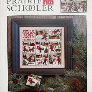 May include: A framed cross-stitch pattern book titled "The Prairie Schooler" with the text "Button Up". The cross-stitch design features winter scenes with people, houses, and trees. The color palette includes red, green, and brown.