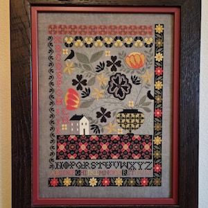May include: Framed cross-stitch sampler with a floral and house design. The artwork features a gray background with colorful flowers, birds, and a house. The frame is dark brown with a red inner border. The sampler includes the alphabet.