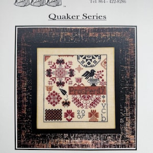 May include: A framed cross-stitch sampler with the word "Prosperity" in the center. The sampler features a variety of geometric and floral patterns in shades of red, black, and cream. The text "Quaker Series" is above the sampler.