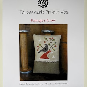 May include: A rectangular cross-stitch pillow featuring a black crow perched on a red Santa hat with green holly branches and red berries. The text reads Threadwork Primitives, Kringle's Crow, Original Designs by Nan Lewis, and an email address.