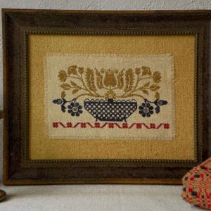 May include: A framed cross-stitch embroidery featuring a central dark blue basket filled with golden yellow floral motifs. Below the basket is a horizontal row of small red cross-shaped patterns. The piece is set against a mustard yellow fabric background.
