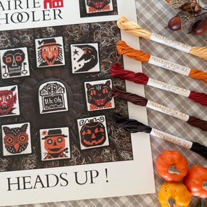 May include: A Halloween-themed cross-stitch pattern with various spooky designs, including a skull, witch, and cat. The image also features spools of silk thread in orange, red, and brown, along with small pumpkins. The text "PRAIRIE SCHOLER" and "HEADS UP!" are visible.