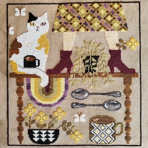 May include: Cross-stitch artwork featuring a calico cat holding a coffee cup, seated on a table with a rainbow arch. The table also displays a plant, spoons, and a patterned mug. The artwork includes a patterned skirt and burgundy boots.