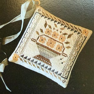 May include: Square, off-white embroidered pillow with a floral design in shades of brown and peach. The pillow features a decorative border and a light blue ribbon for hanging. The design includes a vase with flowers.