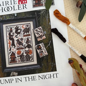May include: A framed cross-stitch pattern titled "Bump in the Night" with Halloween-themed motifs. The design includes witches, owls, and cats. Two skeins of embroidery floss in brown and marigold yellow are also visible.