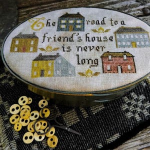 May include: An oval-shaped embroidery hoop containing a cross-stitch sampler with the text "the road to a friend's house is never long" surrounded by four small houses in blue, yellow, and red, accompanied by a pile of light-colored buttons and scissors.
