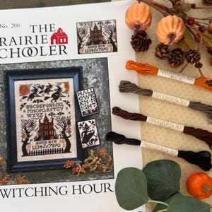 May include: A cross-stitch pattern titled "Witching Hour" by The Prairie Schooler, featuring a spooky house, witches, and the text "Good Bye". Includes orange and brown embroidery floss, pumpkins, and decorative elements.