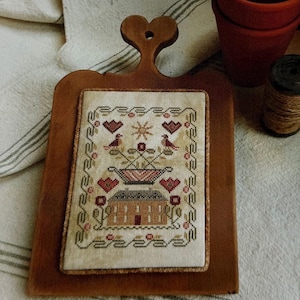 May include: A rectangular wooden board with a heart-shaped handle featuring a mounted cross-stitch sampler. The embroidery displays a house, a flower basket, birds, and hearts in muted red, green, and brown tones on a beige fabric background.