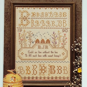 Può includere: Un ricamo a punto croce incorniciato con un tema di api. Il ricamo presenta un alveare, fiori e il testo "Could we live without the bee To fill each hive with sweet honey?" Il ricamo è circondato da una cornice in legno marrone.
