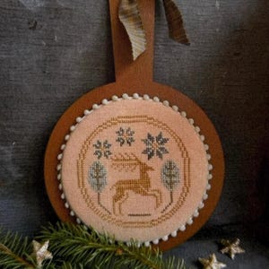 May include: A circular wooden plaque featuring a cross-stitched fabric insert with a light peach background. The embroidery depicts a brown deer, two stylized trees, and three dark blue snowflakes. The edge is trimmed with small white pom-poms.