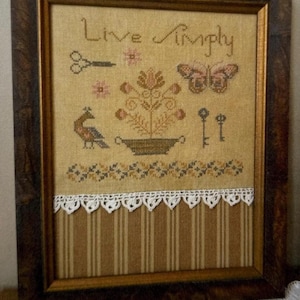 May include: A framed cross-stitch sampler featuring the text Live Simply, a potted flower, a butterfly, a bird, scissors, and two keys. The fabric is tan with a white lace trim separating a lower section of brown and tan vertical stripes.