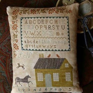 Puede incluir: Una almohada beige con un diseño de punto de cruz que presenta una casa, pájaros y el alfabeto. La casa es amarilla con un techo marrón y una puerta gris. Los pájaros son rojos y están posados en el techo. El alfabeto está en dos filas, con las letras mayúsculas en la fila superior y las letras minúsculas en la fila inferior. Los números del 1 al 9 también están incluidos en el diseño.