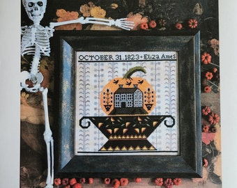 May include: Cross stitch pattern titled "Jack's Urn" featuring a pumpkin with a house inside, surrounded by leaves and a skeleton arm. The pattern is by Kathy Barrick and includes the date "October 31, 1823 - Eliza Ames".