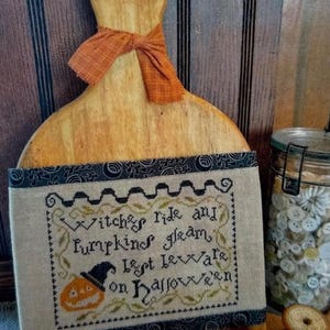 May include: A light-colored wooden cutting board with a handle, adorned with an orange plaid fabric bow. A rectangular piece of beige fabric is attached, featuring black cross-stitched text reading Witches ride and pumpkins gleam, lest beware on Hallowe'en.