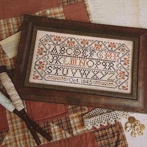 May include: A rectangular framed cross-stitch sampler featuring the alphabet in black and rust-colored thread, with a decorative floral border and the text Oct. 1845. The piece is displayed in a dark brown wooden frame with a visible grain texture.